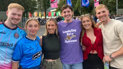 Six people lined up with their arms around each other. There are three boys and three girls. The boys and the girl on the left are wearing blue Gaa shirts, the girl in the middle is wearing a black top, the boy in the middle is wearing a purple Guinness shirt, the girl on the right is wearing a red top, and the boy on the left is wearing a beige shirt.