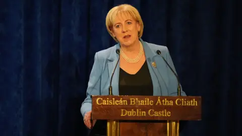 PA Media Heather Humphreys, with short blonde hair, wearing a blue blazer and a black top. She is standing behind a wooden podium with "Caisleán Bhaile Átha Cliath, Dublin Castle" written in gold on the front of it. There is a navy curtain covering the wall behind her. 