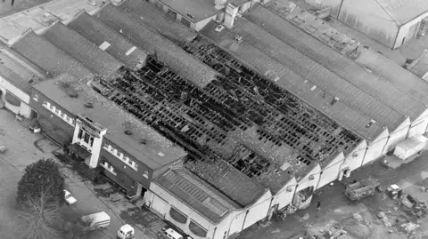 Independent News and Media/Getty Images A black and white aerial shot of Stardust nightclub after the fatal fire. The large, warehouse-like complex consists of several attached buildings with pitched roofs and a main entrance with a flat roof and large windows. More than a third of the pitched roofs are badly burned, with damaged rafters visible.