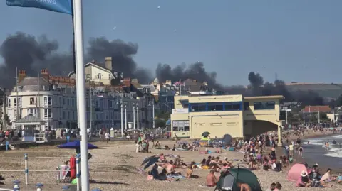 A view of Weymouth beach with crowds of people enjoying the sunshine, with thick black smoke billowing in the background