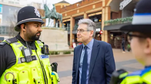 A man in a navy blue suit with a light blue shirt and a navy blue tie is talking to two police officers. He is standing in a city centre with a statue and a large building behind him and signage for The Botanist restaurant.