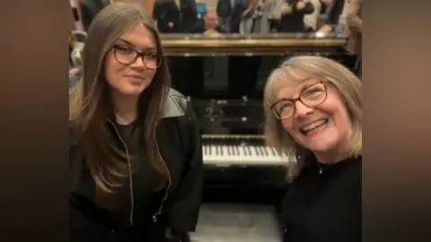 Cambridge University Hospitals Grace Matthews is with her very smiley mother Vicky in front of a large piano. people mill in the background.