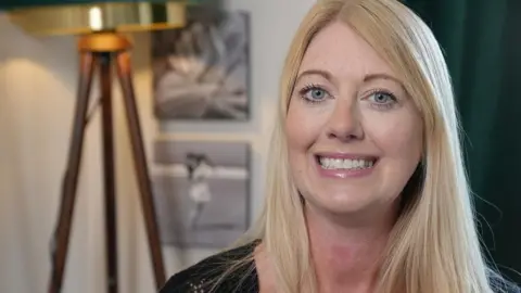 Laura Foster/BBC A woman with long blonde hair is smiling while sat in a living room with a large standing lamp behind her.