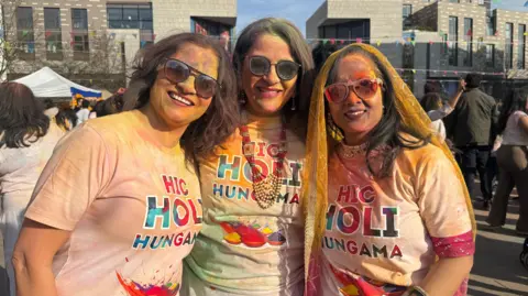 Seema Chugh, Anjali Mavi and Reun Dublish are all wearing white T-shirts which have been covered in coloured powder. They are all stood in line, looking at the camera and smiling. They are all also covered in various coloured powders.