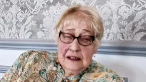 Suffolk Police Joy Middleditch with medium-length light-brown hair, wearing dark brown-framed glasses, and wearing a green, brown, blue and orange top. She appears to have her eyes closed and is leaning back in a chair next to a brown radiator with a silver wallpapered wall behind her.