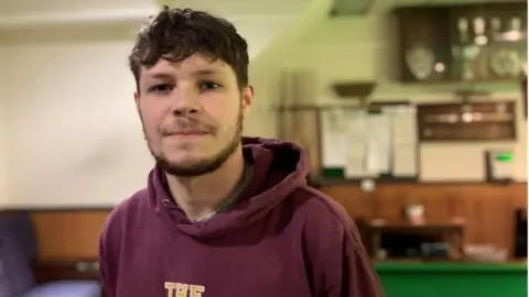 A young man wearing a purple hoodie stands in front of a table and trophy cabinet in a snooker room. He has short, curly brown hair and a bushy brown beard.