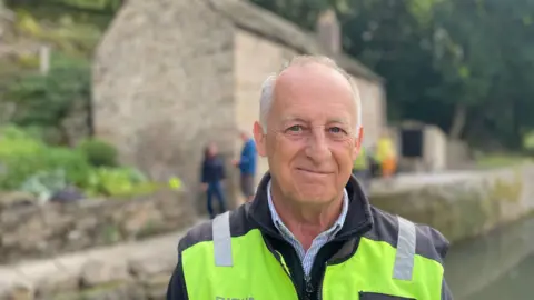 Volunteer project manager Ron Common wearing a hi-vis jacket standing in front of Aqueduct Cottage, a 19th Century lock keeper's cottage with light coloured stone