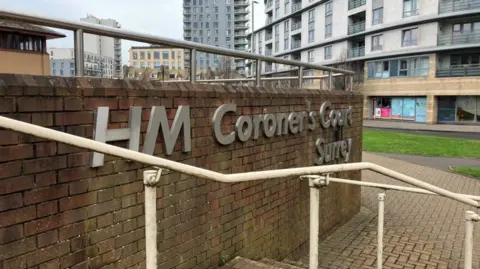 A sign on the side of a brick wall says 'HM Coroner's Court Surrey' with steps leading up to the court and a number of high rise buildings seen in the background.