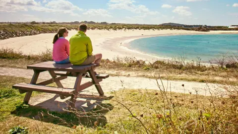 A man and a woman are sitting on the top of a wooden picnic bench on a bright sunny day. He is looking at her and she is looking at the bay in front. The bay has pale sand and a calm blue sea.