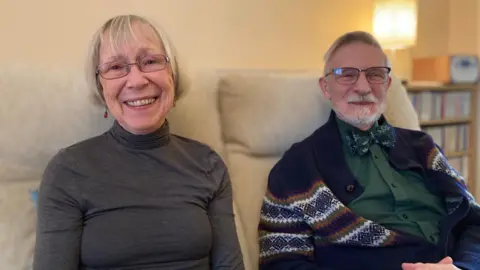 Eve and Peter look into the camera while sitting on a sofa