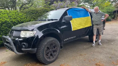 BBC A man in khaki t-shirt and shorts leans on the side of the black Mitsubishi Warrior car, a big 4x4 with a grill on the front, high wheels and a Ukraine flag draped across one of the side windows. It is parked on a road with a bush behind it.