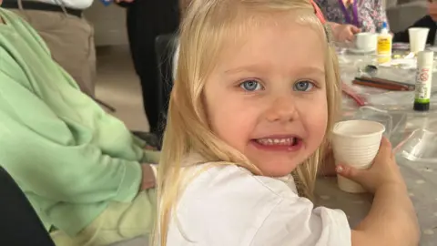 Kate Bradbrook/BBC Indie aged four, wearing a short-sleeved white t-shirt. She is sitting at a plastic covered table, which has glue sticks and pens on it, holding a plastic cup