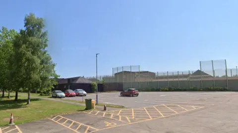 A general view of HMP Huntercombe - which has high metal gates surrounding it - and its car park on a sunny day.