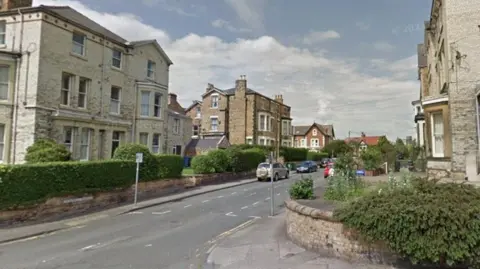 Google Street View image of a residential street lined with tall town houses, with cars parked along the road.