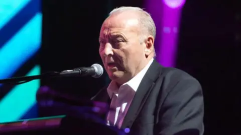 A man with grey hair wearing a dark suit and white shirt playing a keyboard with a microphone in front of him
