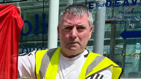 Joao Santos A man wearing a hi-vis jacket over a white top. He is looking at the camera in front of a glass building.