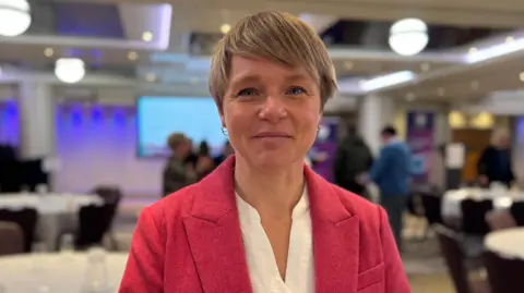 BBC Alice Gill, a woman with short blonde hair and gold earrings. She is wearing a pink blazer over a white blouse. She is looking directly at the camera. Behind her is a conference room, with a large projector screen visible.
