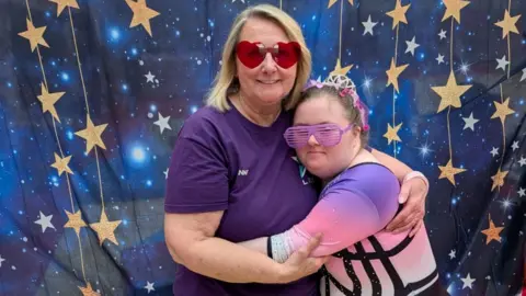 Contributed A mother and daughter embrace and smile at the camera. The mum has short blonde hair and wears a purple T-shirt. The daughter wears purple glasses and a purple and pink gymnastics leotard.