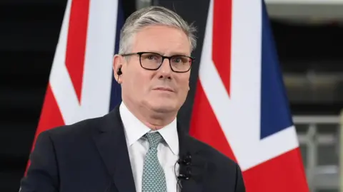 Sir Keir Starmer, dressed in a dark suit and light green tie, stands at a podium speaking. Behind him are two large Union Jack flags. His face is blurred in the image."