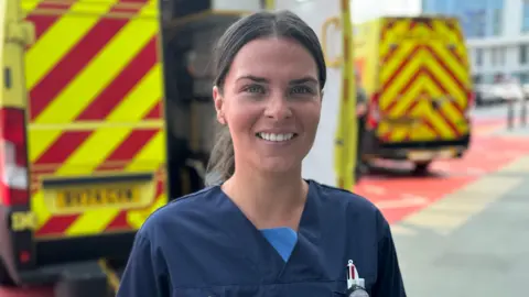 A woman with dark hair pulled behind her head smiles to the camera, showing her teeth. She has dark scrubs top on with a pen in her left pocket. Behind are are the backs of two ambulances with diagonal yellow and red stripes.