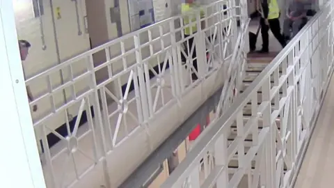 West Yorkshire Police/PA Media Screen grab taken from CCTV showing a prison corridor, with white metal railings and a staircase. Pixelated figures wearing fluorescent uniform can be seen walking towards a partially hidden figure on the left looking at the approaching people.
