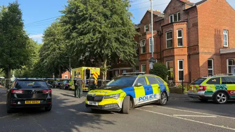 Ambulance vehicles and a police car at the scene of an incident on a tree-lined residential street in Belfast. Four uniformed police officers stand at the open rear door of a bright yellow ambulance. A blue and white police tape cordon stretches across the road towards a terrace of three-storey red brick houses. A blue and white police car is parked sideways across the road in front of the cordon. 