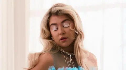 Finn Waring Caity Baser in a promotional shot for her new single. She has long blonde hair worn loose and wears a bright blue tulle dress with a statement gold necklace. She wears red lipstick and white mascara and looks down. She's pictured in a bright, airy room with net curtains covering a sunny window behind her. 