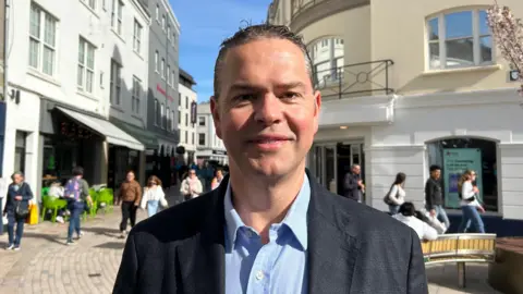 Marcus Calvani, a man with short dark hair, is wearing a light blue collared shirt and a dark blazer. He is staring directly at the camera. He is pictured on a St Helier street, with pedestrians visible behind him.