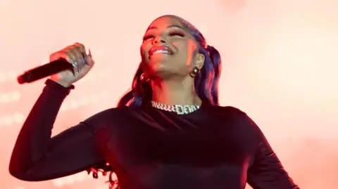 Getty Images Stefflon Don performing at Radio 1's Big Weekend in Luton in May 2024. Stefflon has long hair dyed purple which is tied back in bunches. She wears a long sleeved black top, a large diamond necklace and hoop earrings and smiles while holding a microphone to her face with her right hand 