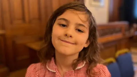 A girl with wavy dark brown hair, wearing a red and white checked school uniform. She is in an old hall with brown panelling behind her, and two blue chairs.
