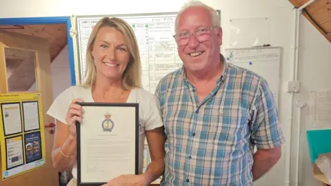 Ann-Marie Dale/Newquay RNLI A picture of a man and a woman stood next to each other smiling at the camera. The woman on the left of the image is wearing a necklace, a white shirt and has blonde hair. She is holding a letter in a frame. The man on the right is wearing glasses, has grey hair and a blue pattern shirt.