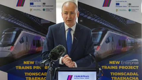 PA Media A balding man in a navy suits standing at a podium. There is a poster with a train on it behind him.