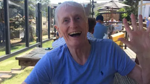 Family handout Melvin Cook. He has short white hair. He is wearing a blue t-shirt. He is pictured outside, holding his hand up to wave at the camera and is smiling, with his mouth open slightly. 