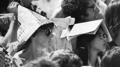A black and white photo of two women, one with a hat made from newspaper on her head sat next to another with an open book perched on her head, watching the Wimbledon championships.
