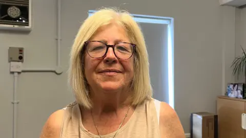 Isabella Verona/BBC An older woman with blonde hair and glasses looks at the camera smiling. She is wearing a beige top with a gold necklace and is standing in front of a white wall.