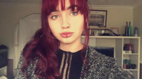 Family handout Christie takes a selfie. She has long thick purple hair and is pouting, with a nose ring and piercing in her bottom lip.