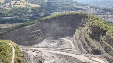 In the foreground is the quarry with lorry tracks and the exposed rock face of the mountain. Beyond that is a view down the valley with trees, fields and villages in the distance.