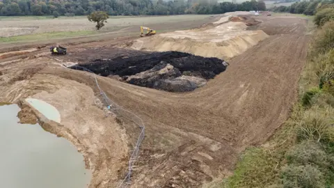 Shaun Whitmore/BBC A part of a field has been dug up, with the middle section of the picture showing a big pile of dirt. At the bottom left of the picture is a big hole filled with water, with a silver wired fence around it. 