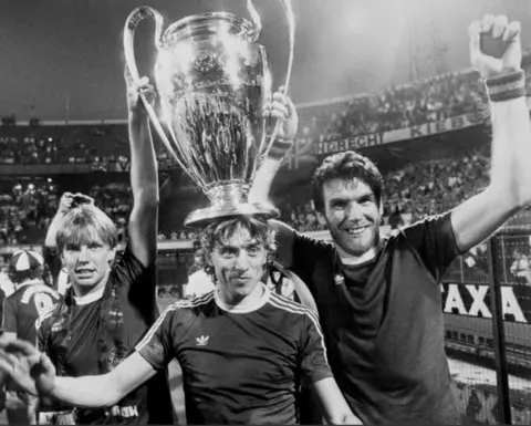 A black and white photo of three smiling footballers, in football shirts, holding a giant trophy aloft. Football crowds can be seen in the background