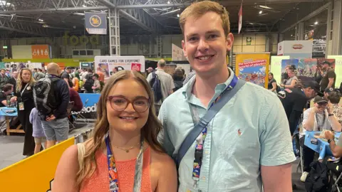 A woman with long blonde hair and an orange dress, smiles as she stands next to a man with a blue shirt. He is also smiling. They are standing in an exhibition hall. People can be seen sitting playing board games and looking at displays.