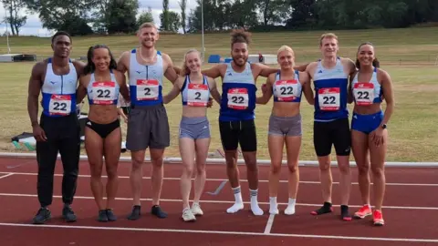 Tom Grantham A group of young athletes huddle together on an outdoor track. They are dressed in blue and white running vests and shorts