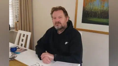 Supplied A man wearing a dark jumper with the white NIKE branding printed on it. He is sat at a table inside. There is a painting hanging on the wall behind him.
