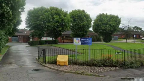 A sign at the entrance says Mayfield School. A large single storey building can be seen and a cat is walking across grass at the front of the school.