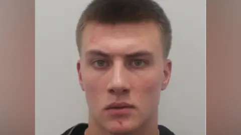 NCA A police mugshot of a man in his early 20s, with short dark blonde hair and a small scratch on his chin, staring into the camera with a serious expression. 