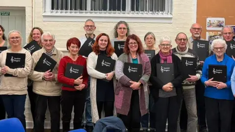 Stotfold Singers Group of people standing indoors in front of a white wall, each holding black folders labeled “Stotfold Singers.” The group is arranged in rows for a posed photo, wearing casual clothing, with a noticeboard and windows visible in the background.