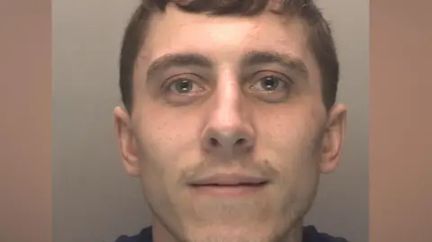 A police mugshot of a man in his early 20s, who has brown hair and light stubble. He is smiling at the camera. 