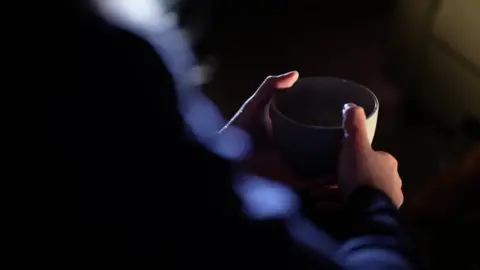 The hands of a woman, sat in the dark, holding a cup.