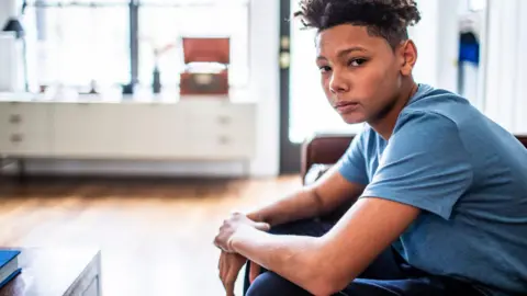 An anxious-looking teenager sits in a room, looking at the camera. 