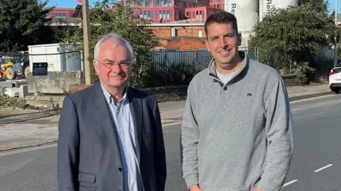 Gloucester City Council Jeremy Hilton and Luke Shervey in front of the site of the former Wessex House in Gloucester. Mr Hilton is wearing a grey blazer and blue striped shirt. He has short grey hair and glasses. Mr Shervey is standing next to him. He has short dark hair and is wearing a grey jumper and black jeans. Both men are looking at the camera and smiling. Behind them, cranes and building equipment and vehicles can be seen. 