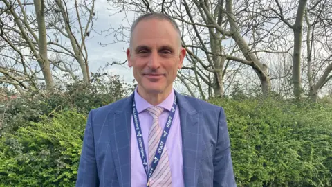 Mark Bolton is smiling at the camera. He is a wearing a suit and tie. His suit is blue and he is wearing a lilac coloured shirt. He also has a blue lanyard. He is standing in front of a hedge and trees.